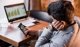 This is a man who has placed his left hand on the side of his face. He is using a mobile phone, and in front of him on the table, there is an open laptop